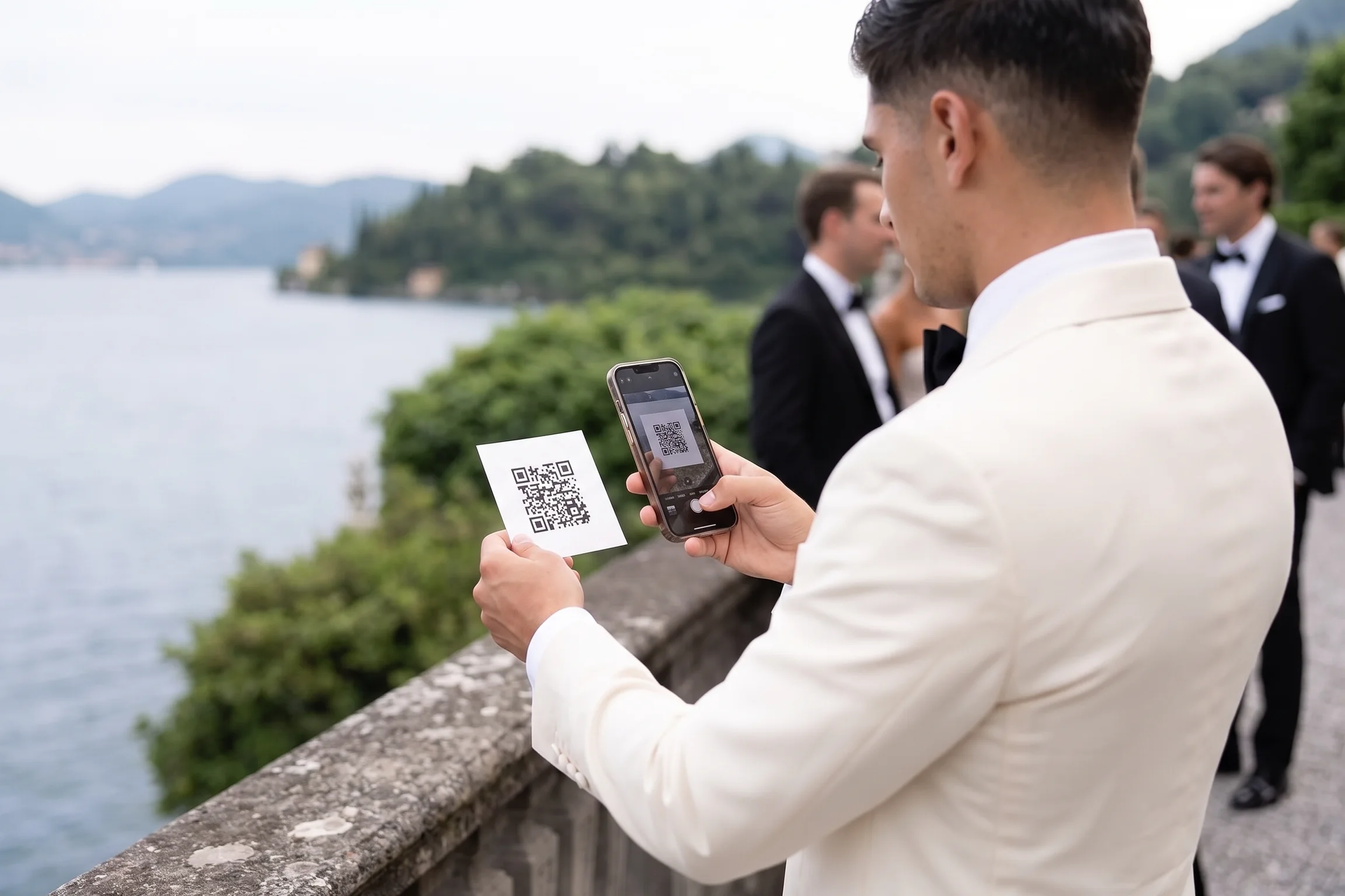 Wedding guest in an ivory dinner jacket scanning the gallery QR card with his phone at a lakeside balustrade